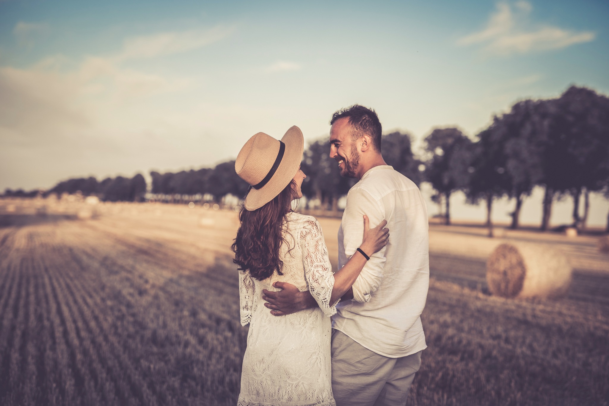 Para zakochanych, on obejmuje ją w talii, ona w kapeluszu, na tle pola ze słomianymi balotami, w ciepłym świetle.