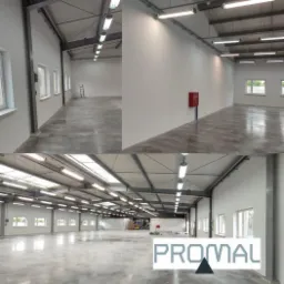 Wnętrze hali magazynowej z świeżo pomalowanymi ścianami na biało i betonową posadzką, widoczne okna i oświetlenie sufitowe, w tle drabina i regały magazynowe, w rogu ściany czerwona skrzynka...