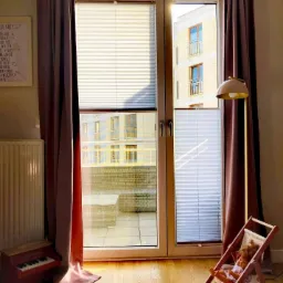 Pokój dziecięcy z widokiem na balkon przez okno z podwójnymi plisowanymi roletami w kolorze białym, zasłoniętymi po bokach brązowymi zasłonami, z zabawkami na podłodze i lampą stojącą.