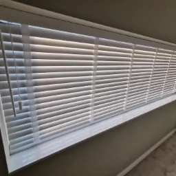 Zamknięte, białe żaluzje poziome w oknie, z widocznym sznurkiem do regulacji i odbiciem światła na parapecie.