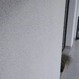 Biała ściana zewnętrzna z fakturą baranka, widoczny fragment okna i doniczka z trawą ozdobną