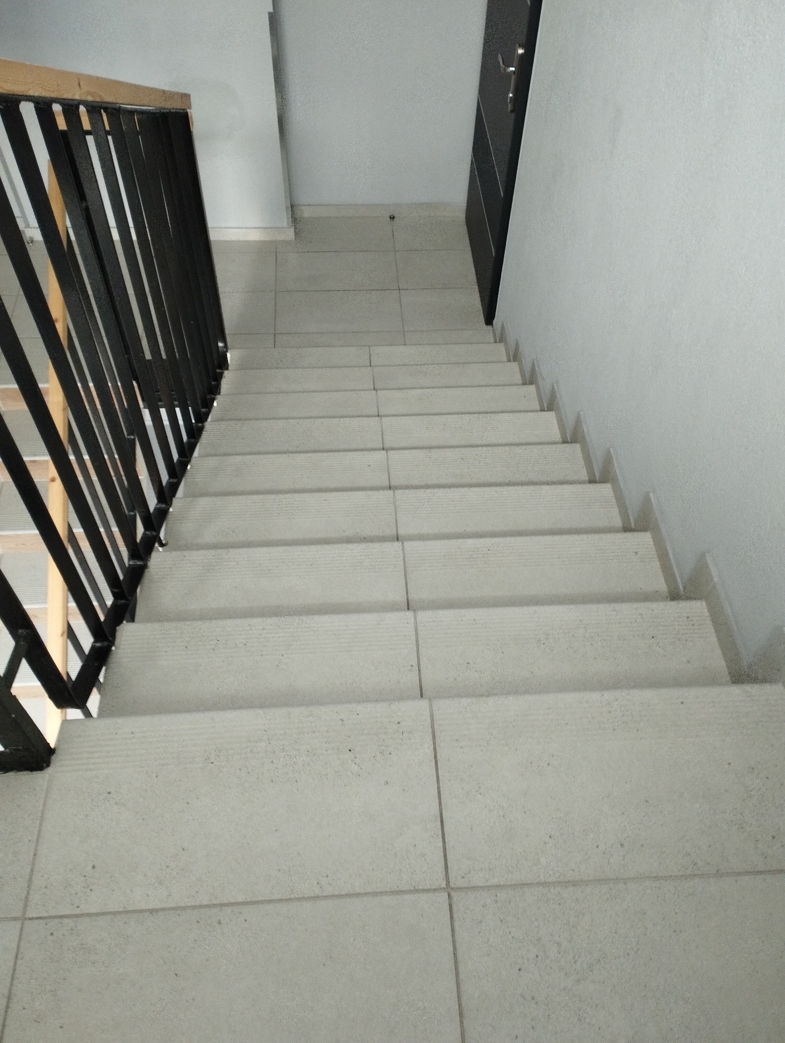 Schody wyłożone jasnymi, kwadratowymi płytkami ceramicznymi, z czarną metalową balustradą po lewej stronie i czarnymi drzwiami na górze schodów.