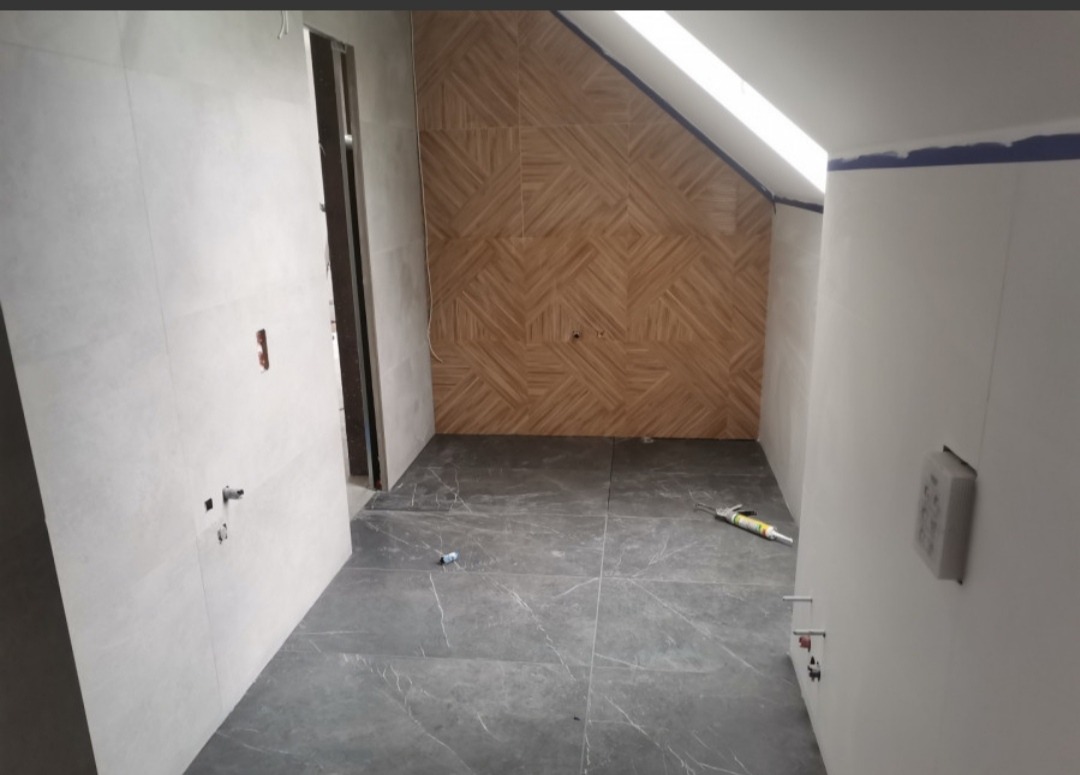 Łazienka w trakcie prac: ułożona podłoga z szarych płytek imitujących marmur, ściana wyłożona drewnopodobnymi płytkami w geometryczny wzór, widoczny pistolet do silikonu i nieukończona ściana...