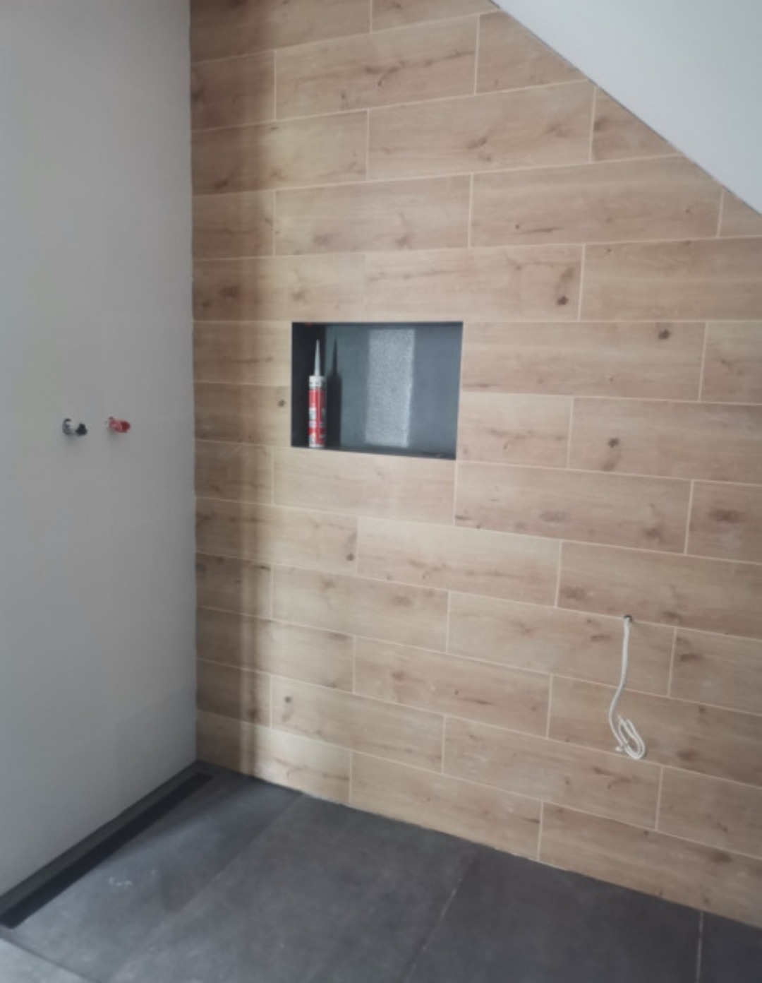 Wykończona ściana łazienki z płytek imitujących drewno, wnęka z ciemnoszarą okładziną, tuba silikonu wewnątrz wnęki, szara podłoga, widoczne przyłącza wodne na ścianie.