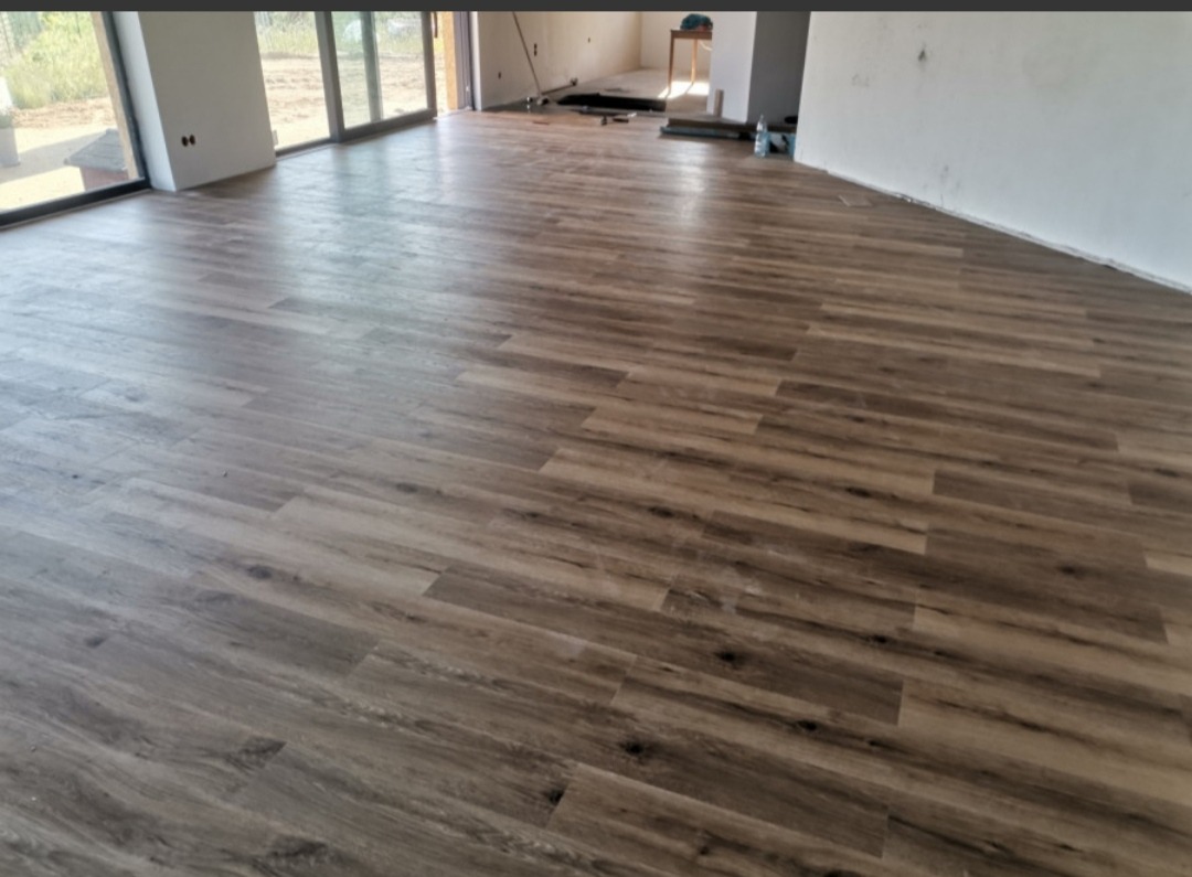 Ułożone panele podłogowe imitujące drewno w przestronnym, jasnym pomieszczeniu z dużymi oknami; widoczne niedoskonałości na ścianach i brak listew przypodłogowych.