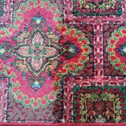 Fragment wzorzystego, kolorowego dywanu z dominującą czerwienią, różem i zielenią, z wyraźną fakturą włókien i motywami roślinnymi w stylu orientalnym.