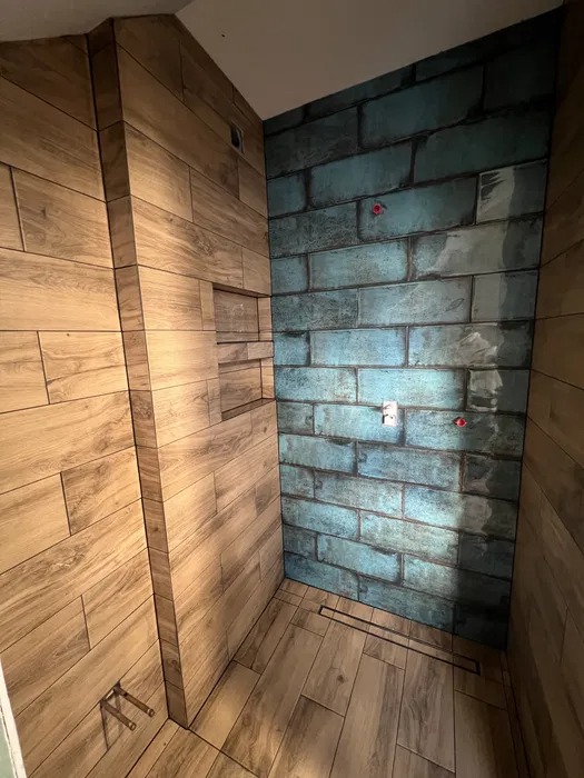 Wykończona kabina prysznicowa z drewnopodobnymi płytkami na ścianach i podłodze, oraz turkusowymi płytkami imitującymi cegłę na jednej ze ścian, widoczne rury instalacji wodnej i odpływ liniowy.