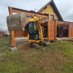 Żółta mini koparka JCB 8018 CTS na trawniku przy drewnianej pergoli, z łyżką uniesioną nad przygotowywanym fundamentem z kamieni
