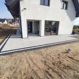 Usługi remontowo budowlane Krzysztof Kasprusz - Nowoczesny taras z jasnoszarej kostki brukowej z czarnym obramowaniem, przylegający do białego domu z dużymi oknami przesuwnymi, widoczny pracownik przy krawędzi tarasu i przygotowane kostki obok.