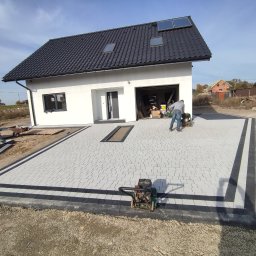 Usługi remontowo budowlane Krzysztof Kasprusz - Świeżo ułożona, jasnoszara kostka brukowa z czarną obwódką przed nowoczesnym, białym domem z czarnym dachem; mężczyzna zagęszcza kostkę zagęszczarką wibracyjną, widoczne narzędzia i materiały...