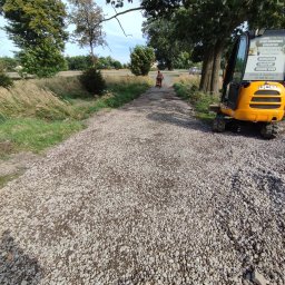 Usługi remontowo budowlane Krzysztof Kasprusz - Żwirowa droga gruntowa w trakcie tworzenia, ubijana zagęszczarką przez pracownika bez koszulki, obok stoi żółta minikoparka JCB z naklejką reklamową usług brukarskich i transportowych.