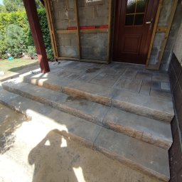 Usługi remontowo budowlane Krzysztof Kasprusz - Wykończone schody wejściowe z kostki brukowej w Orzeszu, prowadzące do drewnianych drzwi, z widocznymi elementami konstrukcji ściany i częściowo ułożoną kostką przed wejściem.