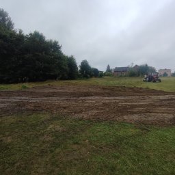 Usługi remontowo budowlane Krzysztof Kasprusz - Teren wyrównany ziemią, z widocznymi śladami opon, w tle drzewa i budynki, pochmurne niebo.