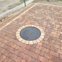 Usługi remontowo budowlane Krzysztof Kasprusz - Nowo ułożona, czerwono-brązowa kostka brukowa z dekoracyjnym obramowaniem z jaśniejszych kostek oraz okrągłą studzienką kanalizacyjną na środku, widoczny fragment ogrodzenia panelowego.