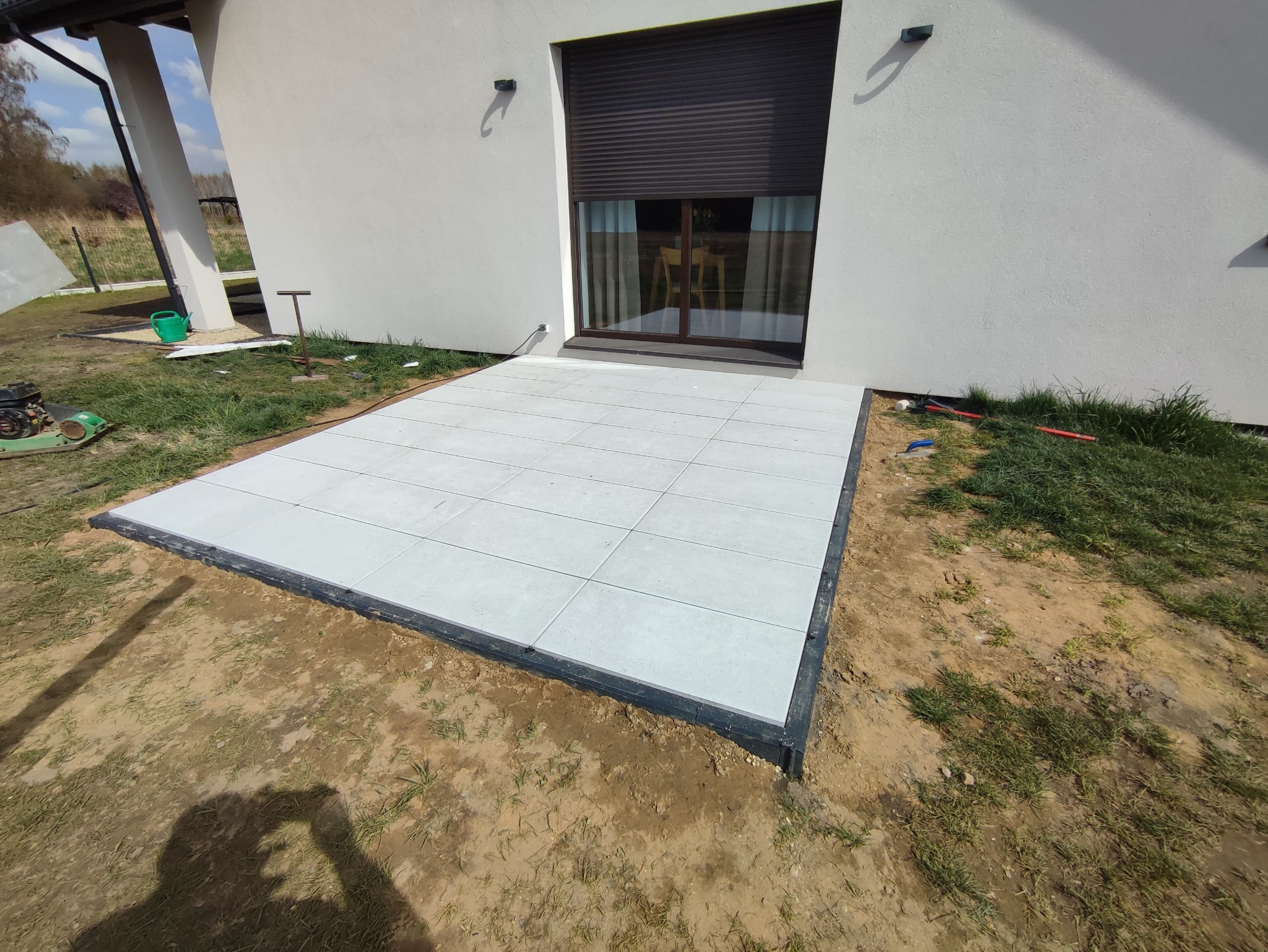 Nowo ułożony taras z jasnoszarej kostki brukowej przy białym domu z brązowymi roletami, widoczny zagęszczarka w tle, teren wokół jeszcze niezagospodarowany.