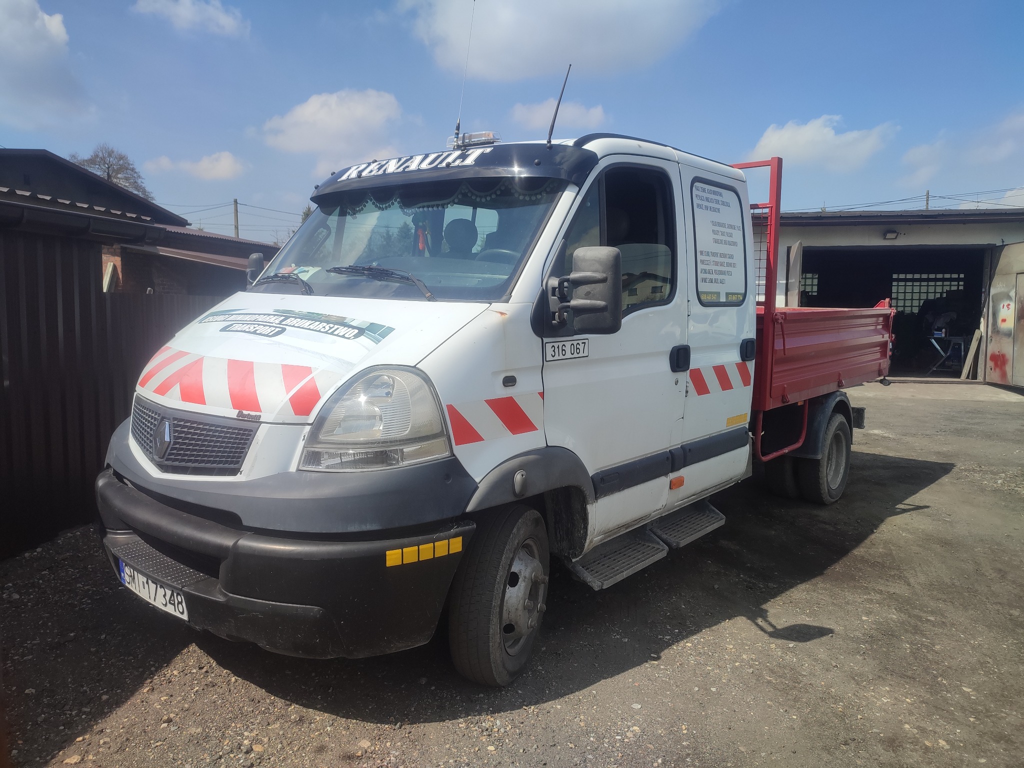 Biały samochód dostawczy Renault z czerwoną skrzynią ładunkową, oznakowany pasami ostrzegawczymi, zaparkowany na żwirowym podłożu przed garażem.