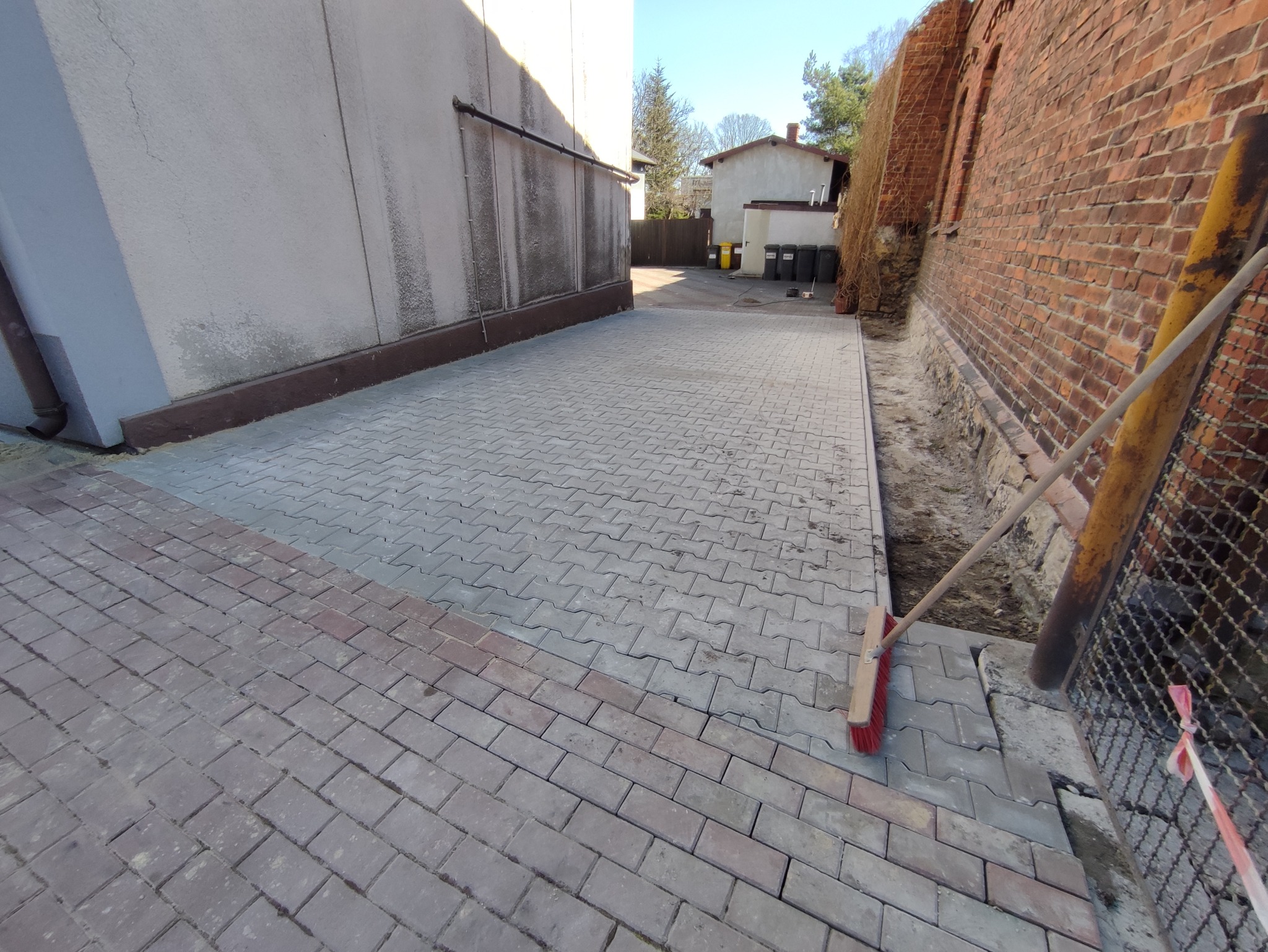 Nowo ułożona szara kostka brukowa w kształcie litery H, obrzeża z czerwonej kostki, szczotka do zamiatania oparta o ceglany mur, widok na budynki w tle.