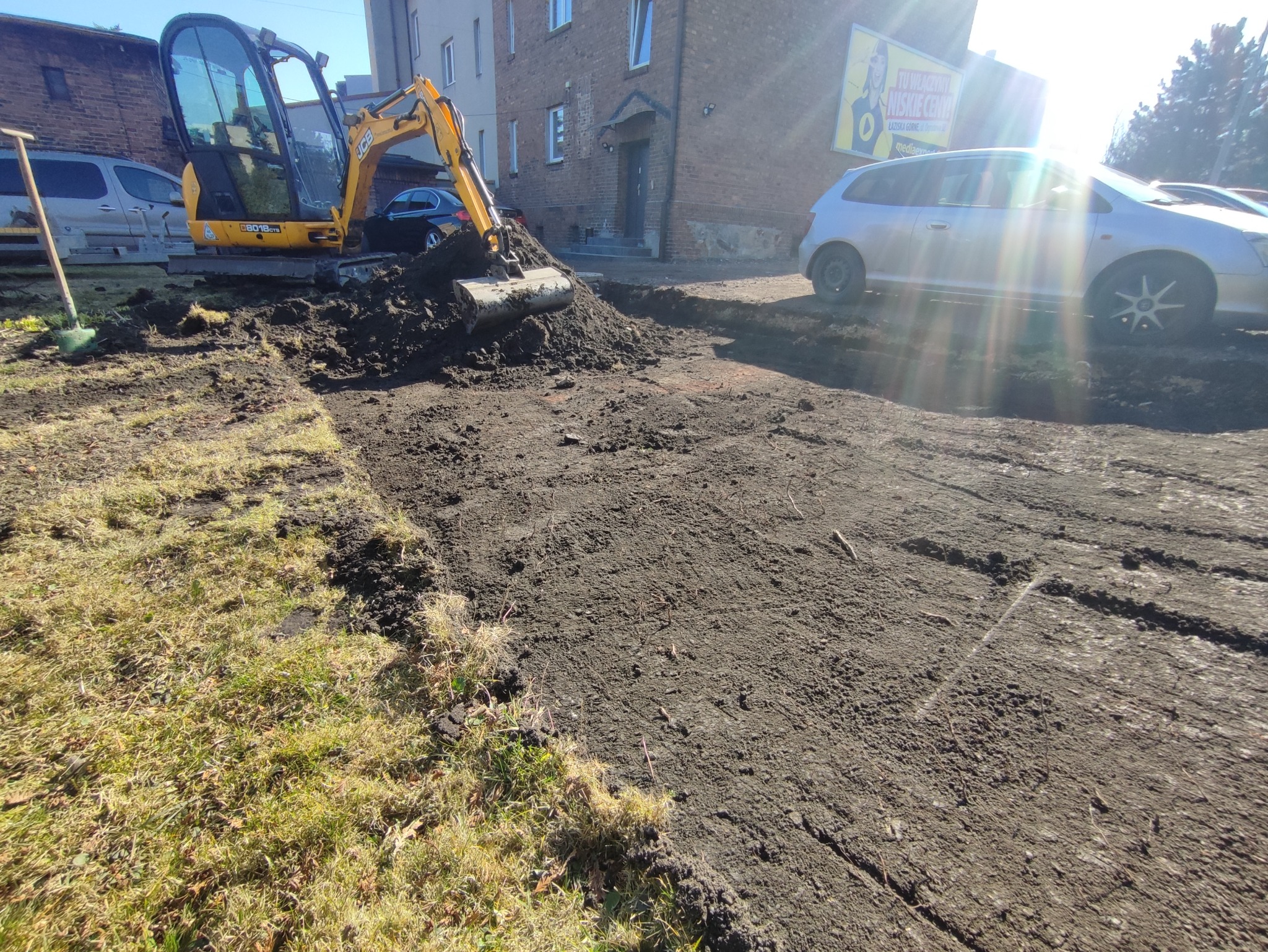 Żółta mini koparka JCB wyrównuje teren pod drogę, widoczne ślady opon samochodowych i budynek w tle, przygotowanie podłoża.