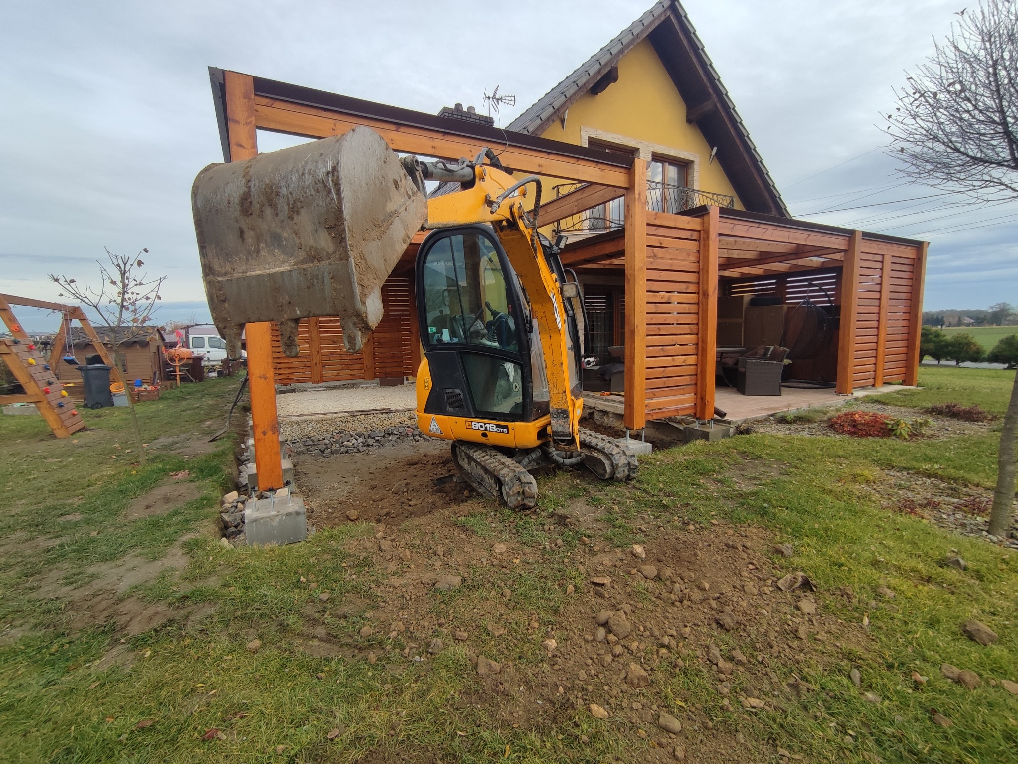 Żółta mini koparka JCB 8018 CTS na trawniku przy drewnianej pergoli, z łyżką uniesioną nad przygotowywanym fundamentem z kamieni