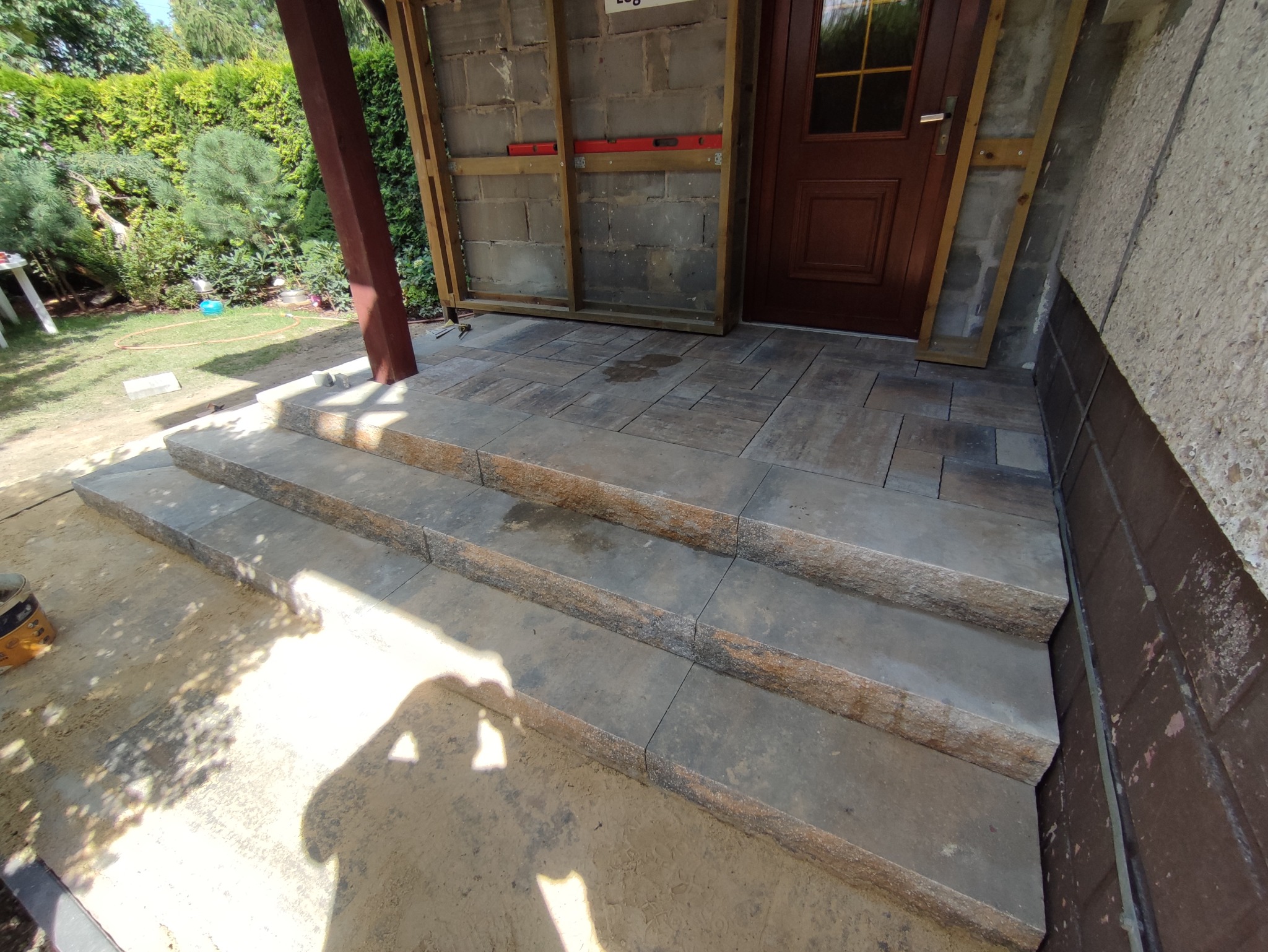 Wykończone schody wejściowe z kostki brukowej w Orzeszu, prowadzące do drewnianych drzwi, z widocznymi elementami konstrukcji ściany i częściowo ułożoną kostką przed wejściem.