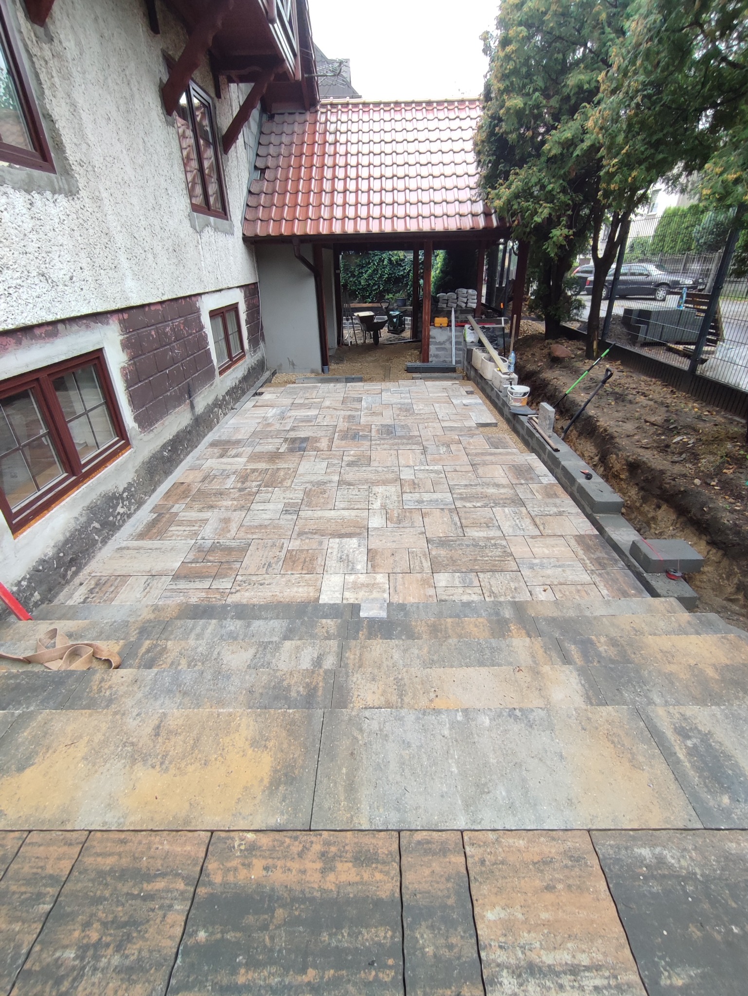 Nowo ułożona kostka brukowa w odcieniach brązu i szarości, prowadząca do domu z czerwonym dachem, widoczne narzędzia i przygotowane materiały budowlane w tle.