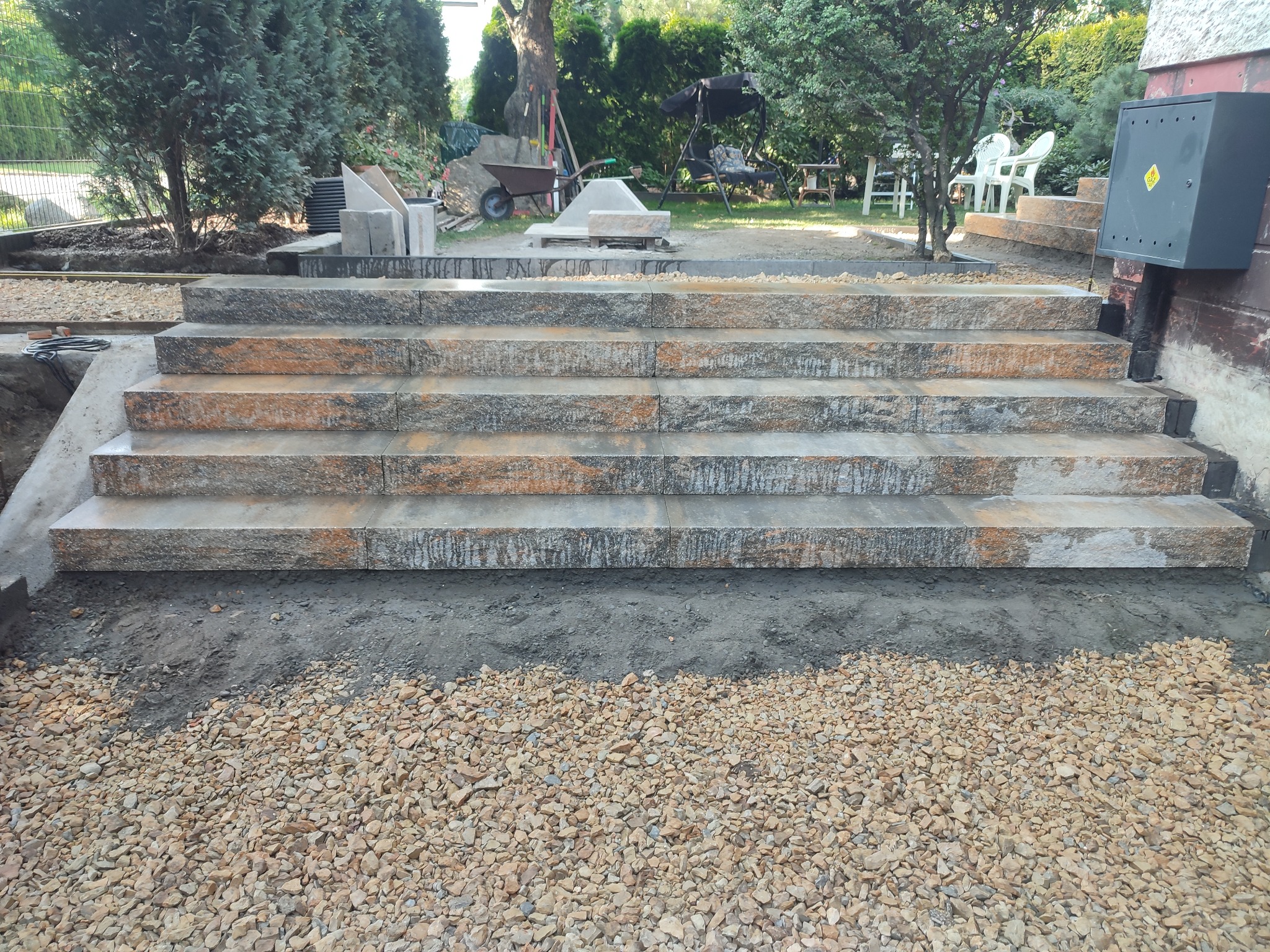 Kamienne schody zewnętrzne z widocznymi śladami montażu, podsypka żwirowa u podstawy, w tle zieleń ogrodu i elementy wyposażenia.