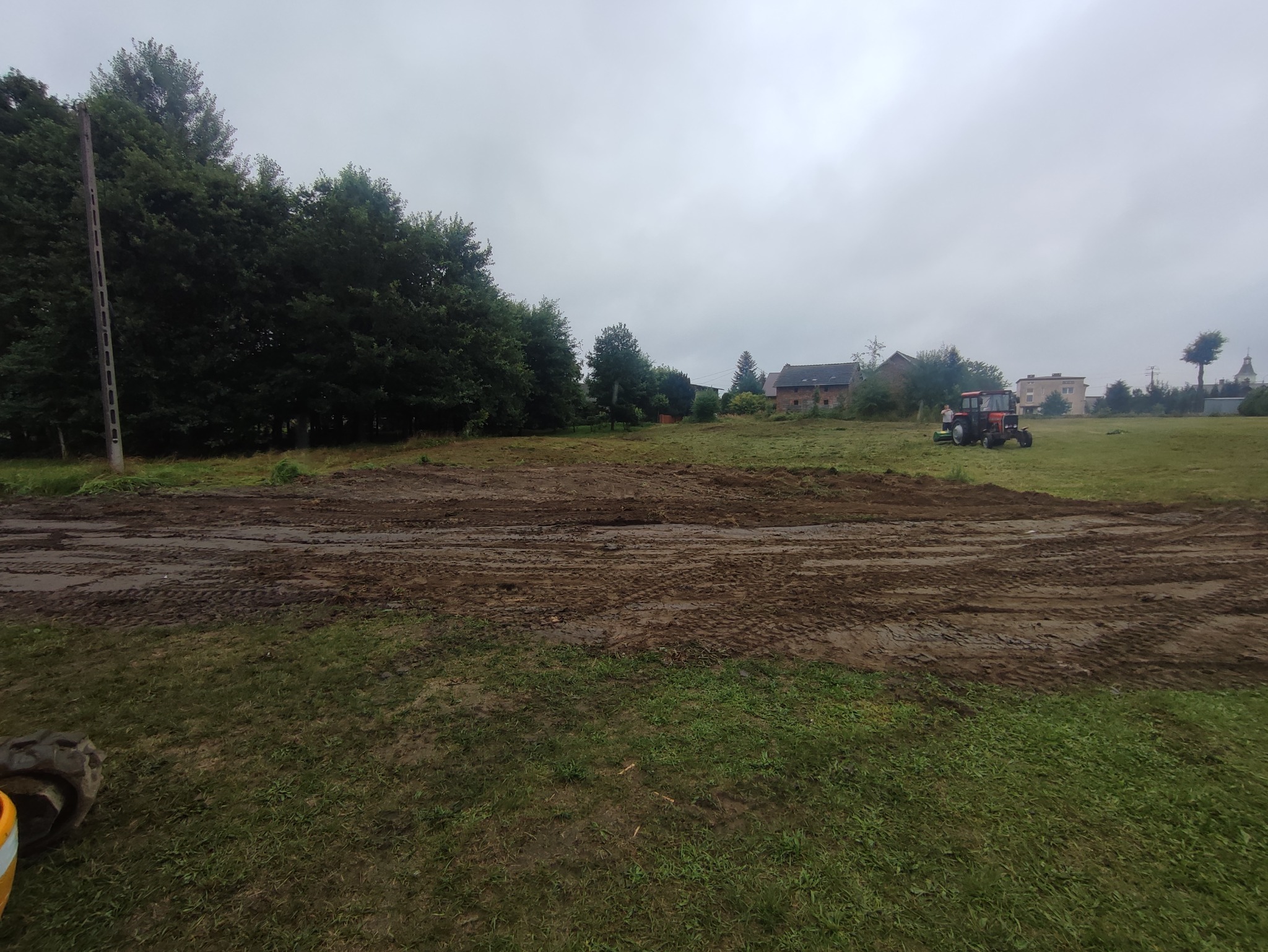 Teren wyrównany ziemią, z widocznymi śladami opon, w tle drzewa i budynki, pochmurne niebo.