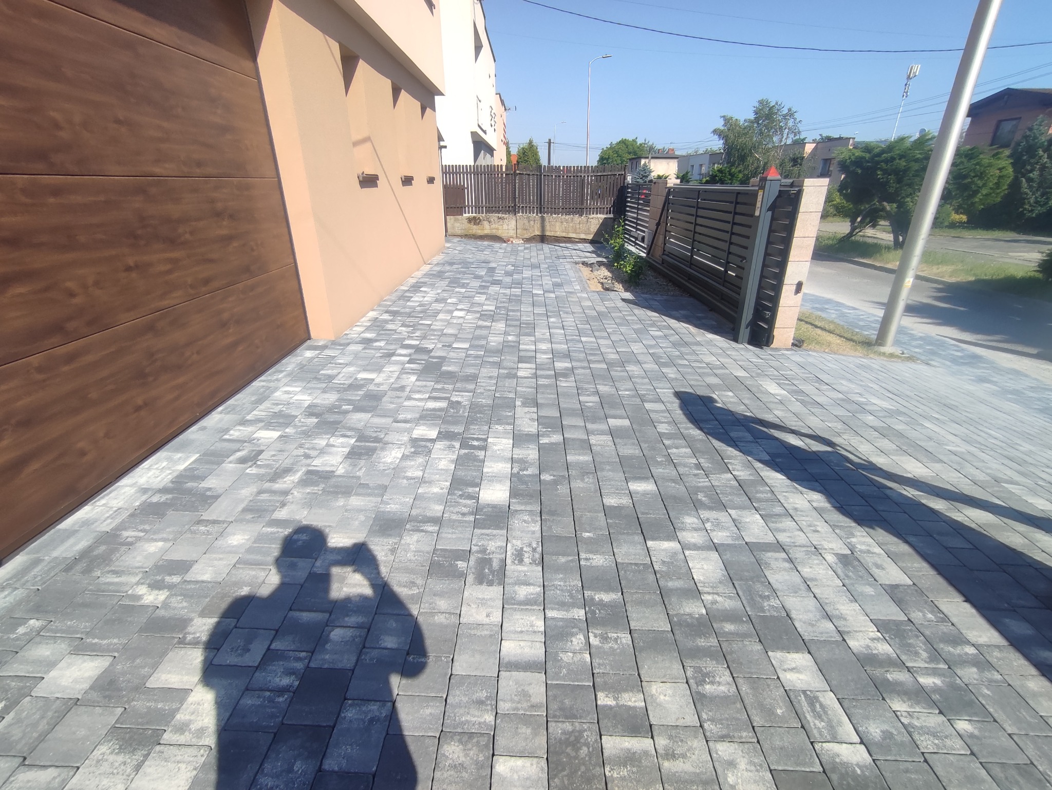 Ułożona szara kostka brukowa na podjeździe, widoczny cień osoby robiącej zdjęcie, brama garażowa w kolorze drewna, ogrodzenie posesji w tle.