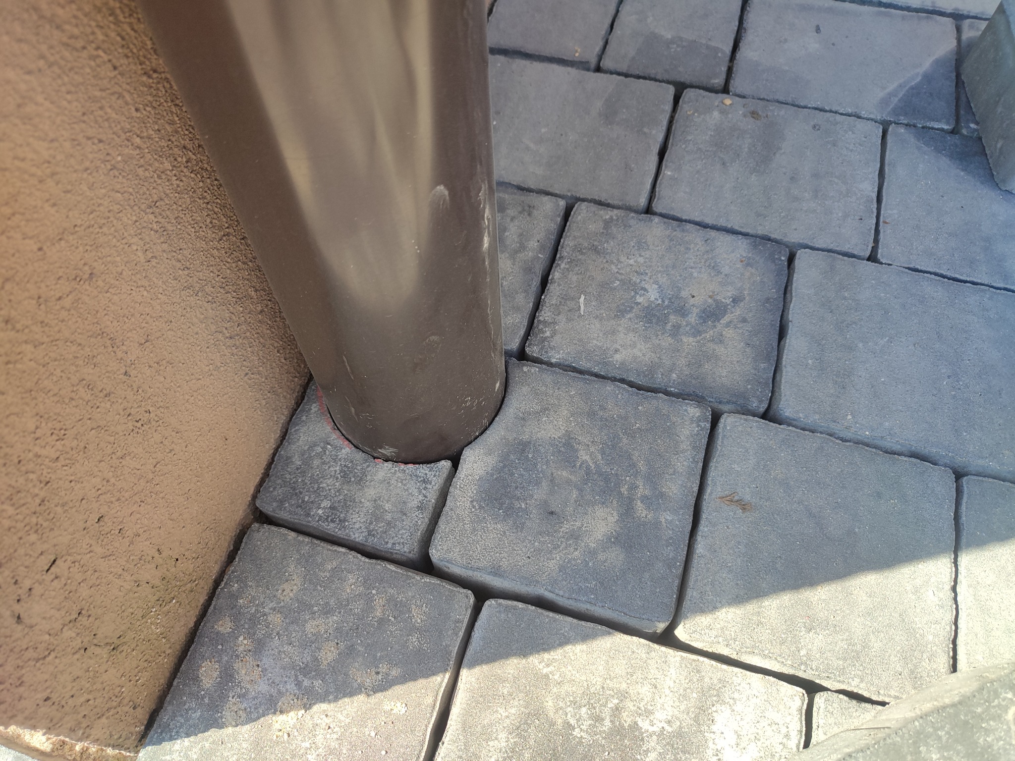 Szare kostki brukowe ułożone wokół brązowej rury spustowej przy ścianie budynku. Krawędzie kostek są lekko zaokrąglone, a na powierzchni widoczne są drobne zarysowania. Część kostek oświetlona jest...