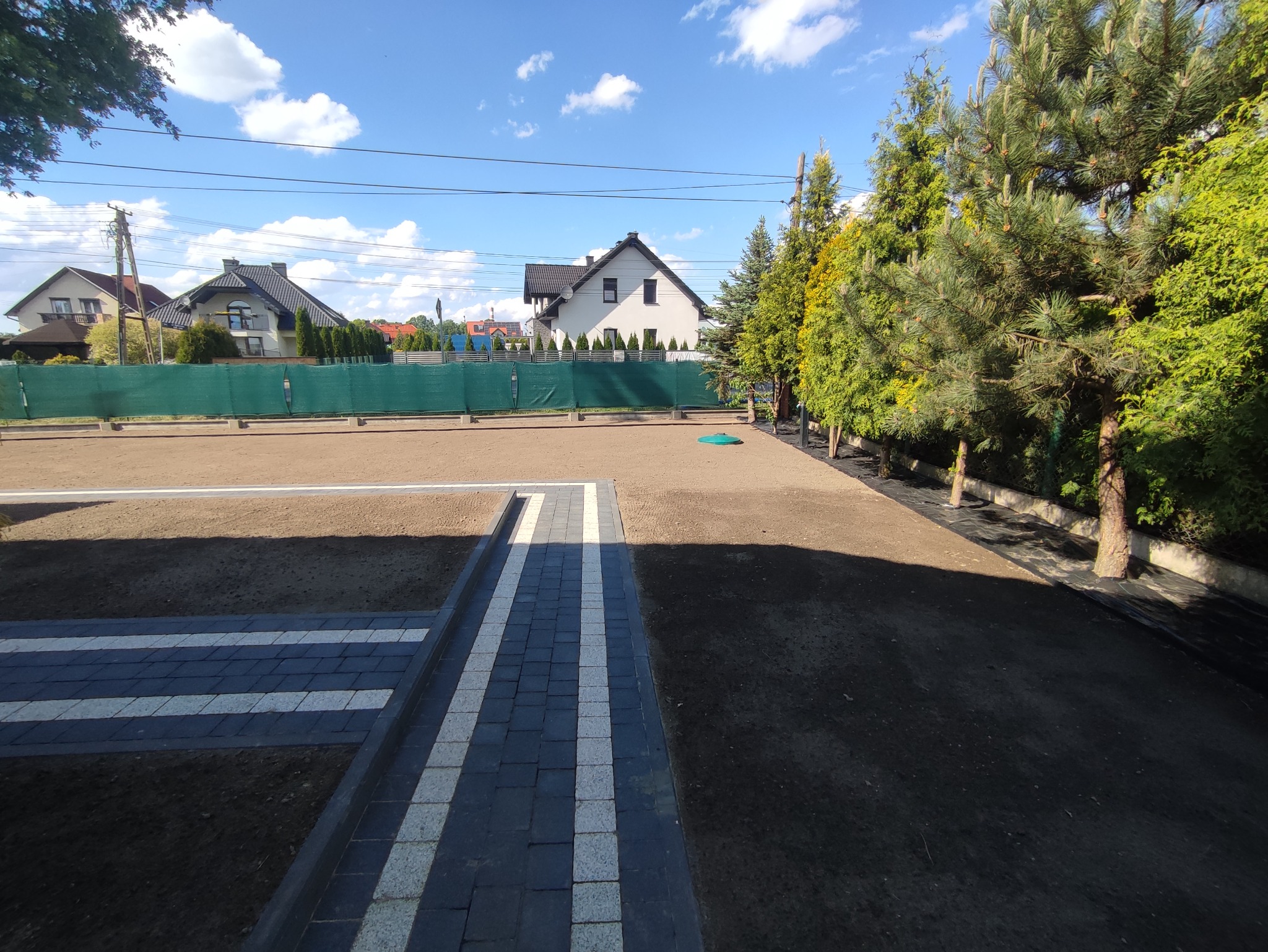 Przygotowany teren pod trawnik z rolki, widoczny fragment ścieżki z kostki brukowej w kolorze szaro-białym, w tle ogrodzenie z siatki maskującej i zabudowa jednorodzinna.