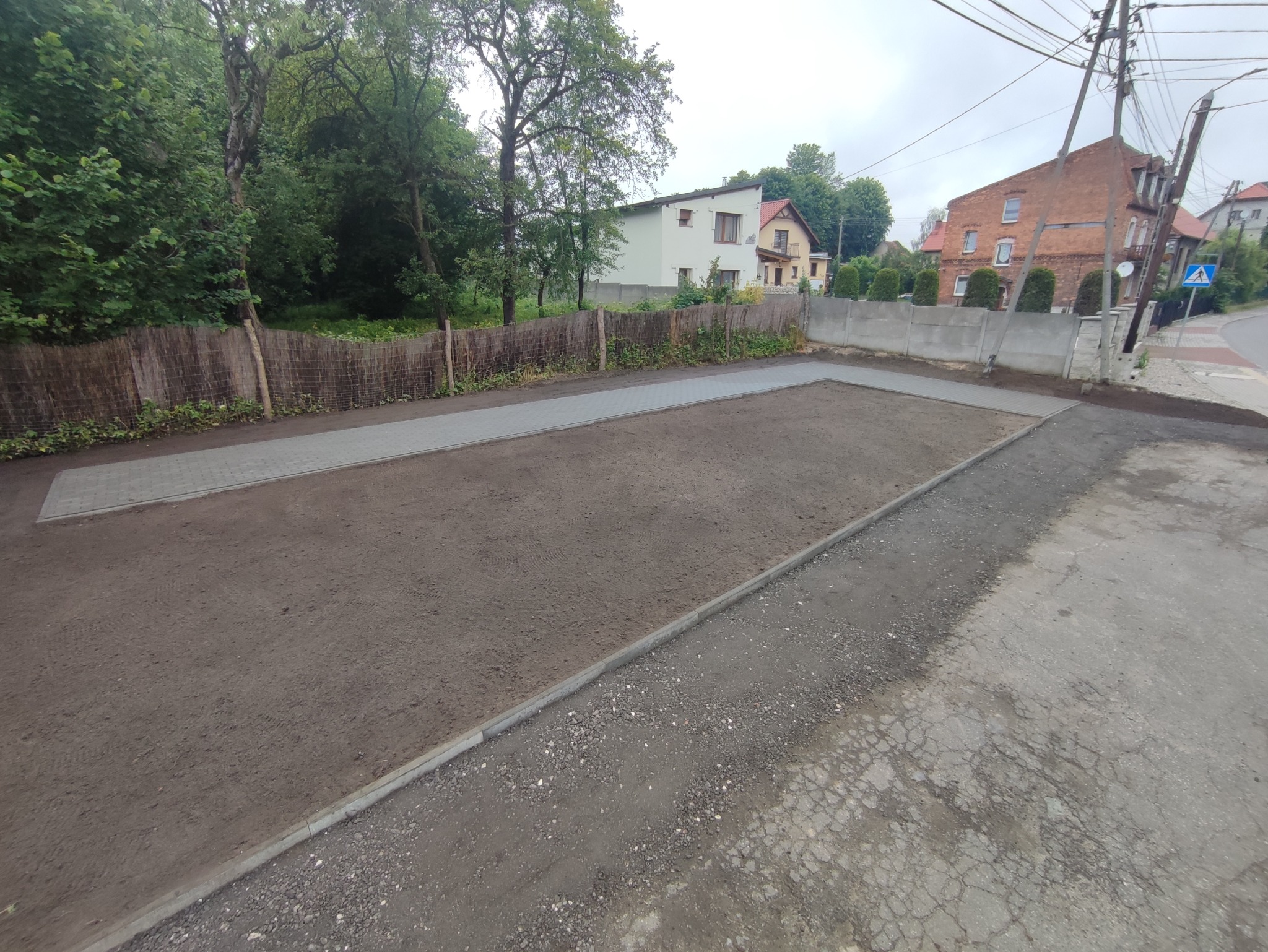 Przygotowanie terenu pod kostkę brukową: wyrównana powierzchnia ziemi z obrzeżami z kostki, widoczne fragmenty ułożonej kostki w tle, budynki mieszkalne w oddali, pochmurne niebo.