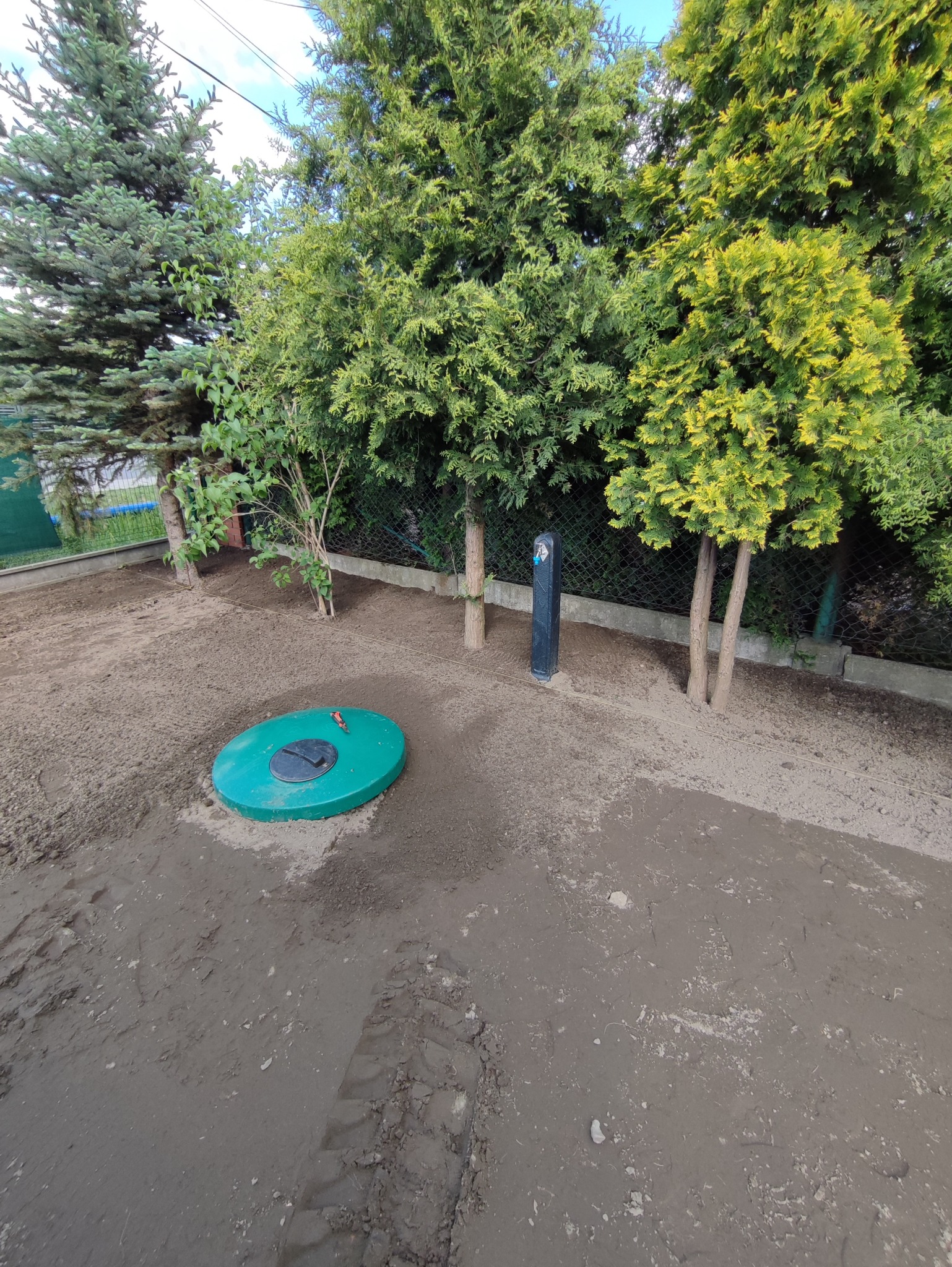 Zielona studzienka kanalizacyjna z czarną pokrywą, otoczona świeżo wyrównanym piaskiem i młodymi drzewami, widoczne ślady opon na błotnistym podłożu.