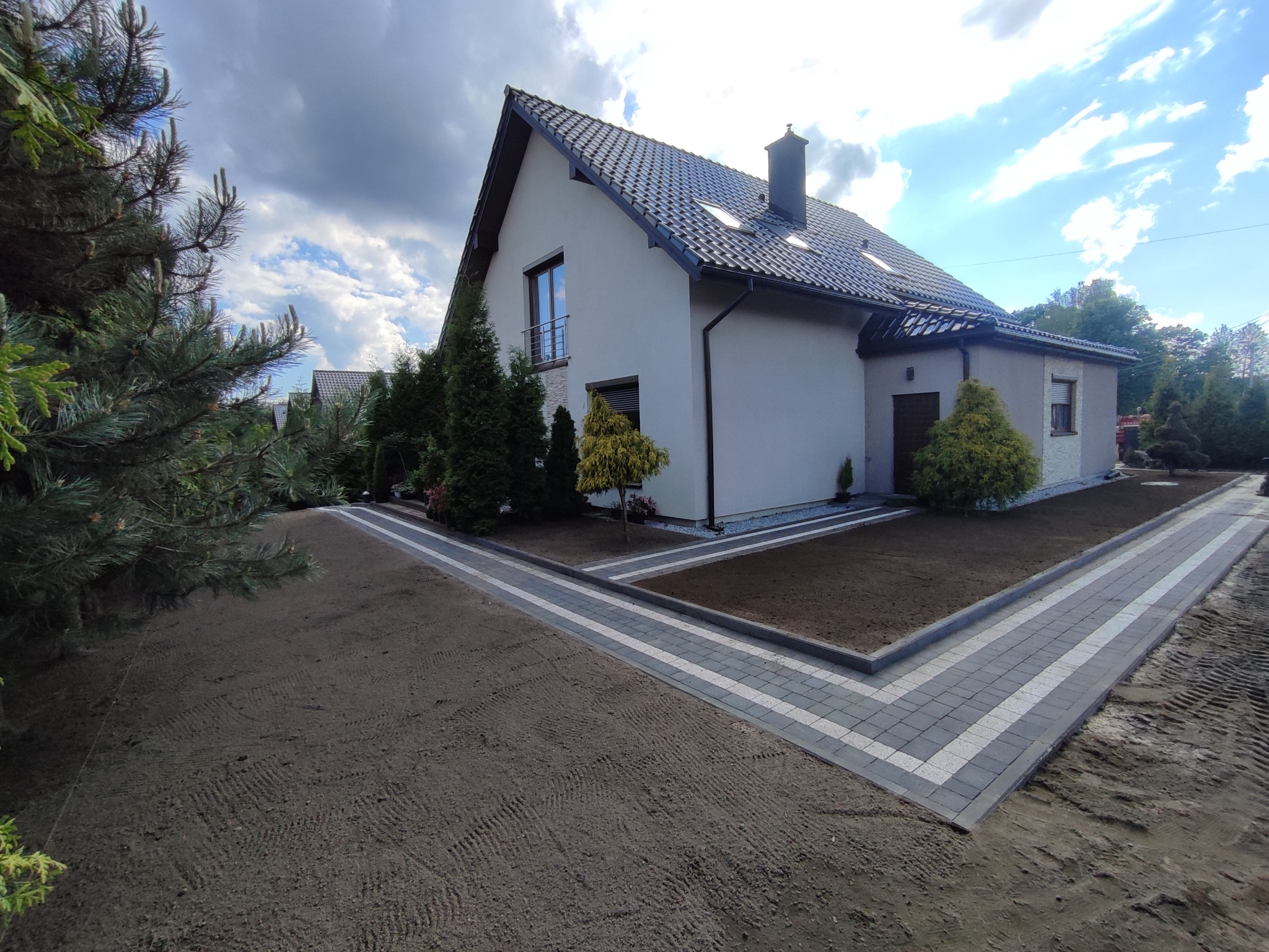 Nowo ułożona, dwukolorowa kostka brukowa tworząca ścieżkę wokół domu z przygotowanym podłożem pod trawnik i posadzone drzewka iglaste.