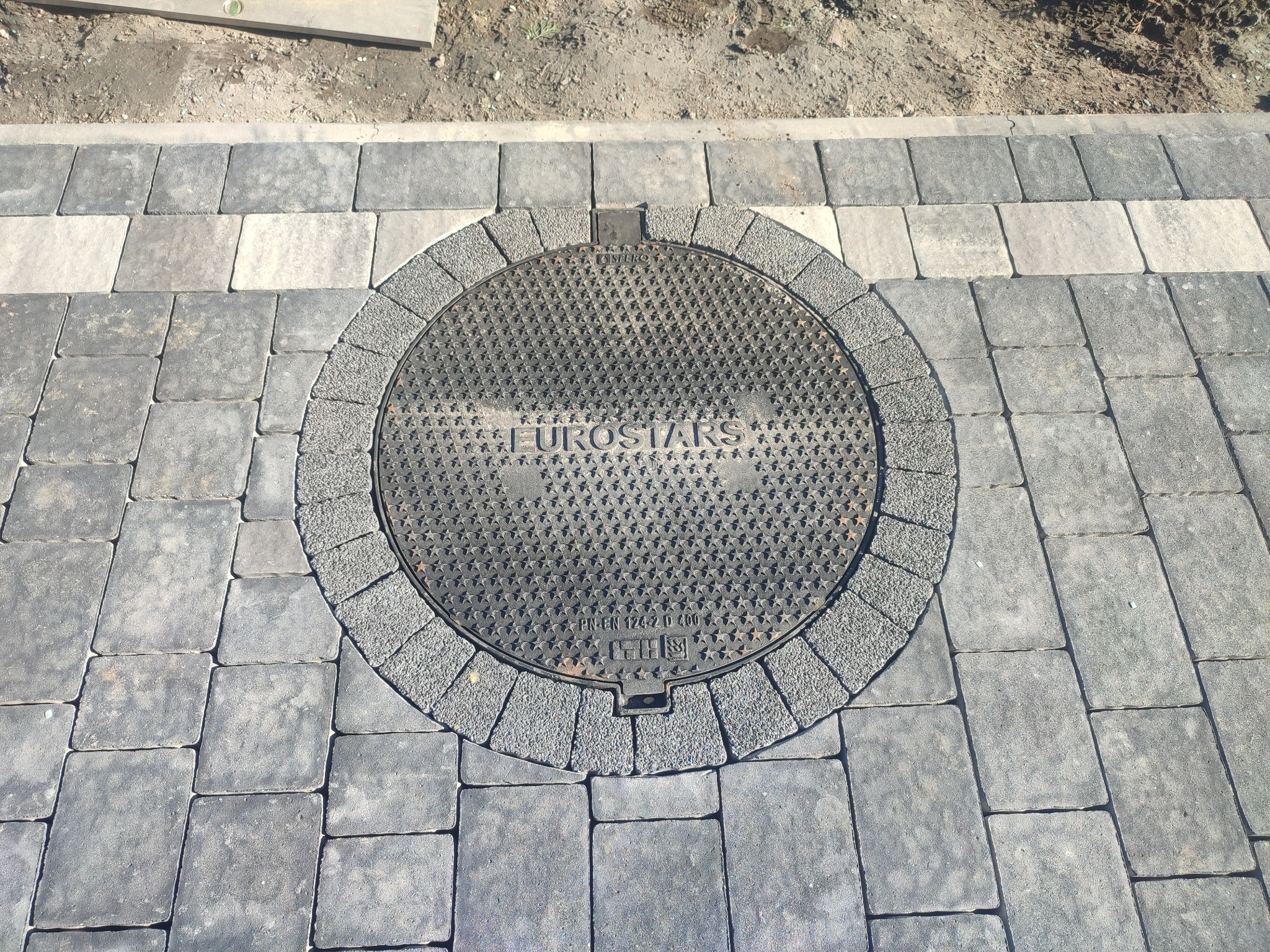Szara kostka brukowa ułożona wokół okrągłej studzienki kanalizacyjnej z napisem EUROSTARS, widok z góry.
