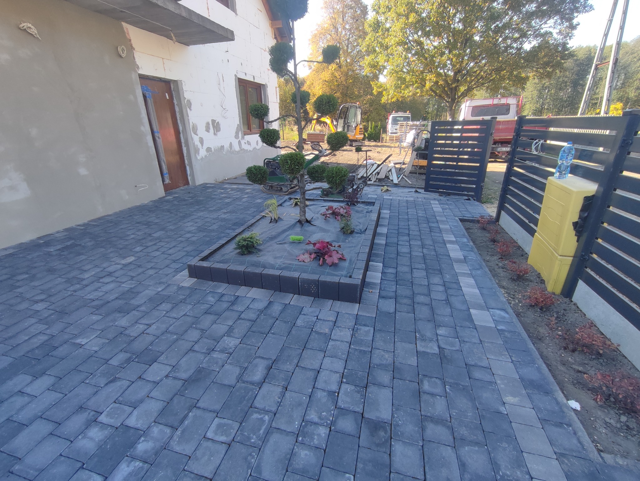 Ułożona szara kostka brukowa z centralnie umieszczonym rabatą z ozdobnym drzewkiem i roślinami, częściowo przykrytą siatką, na posesji z widocznym budynkiem w trakcie wykończenia i sprzętem...