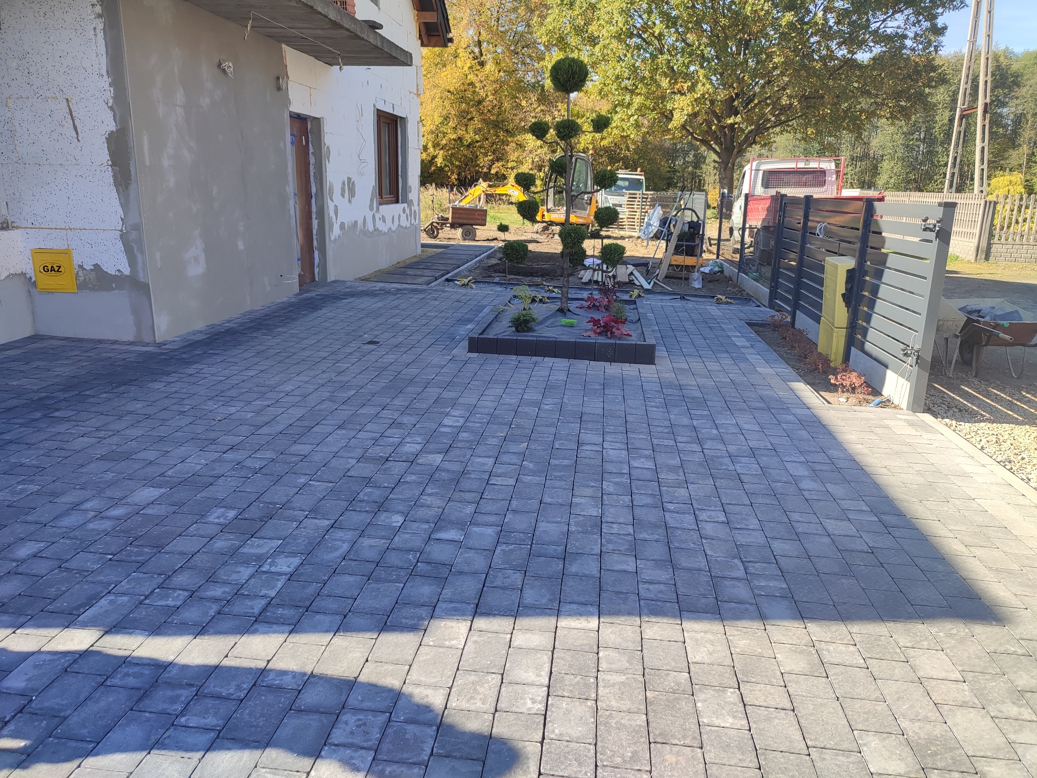 Ułożona szara kostka brukowa na podjeździe domu z małym klombem z drzewkiem bonsai, w tle sprzęt budowlany i ogrodzenie.