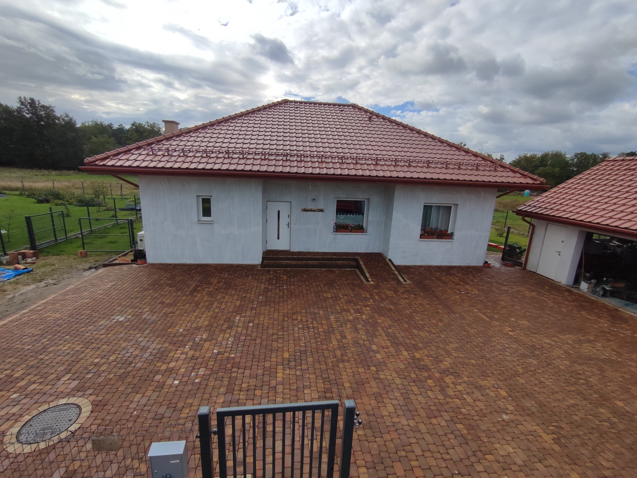 Szeroki widok na dom z czerwonym dachem i podjazd z kostki brukowej w odcieniach brązu i beżu, widoczne elementy ogrodzenia.