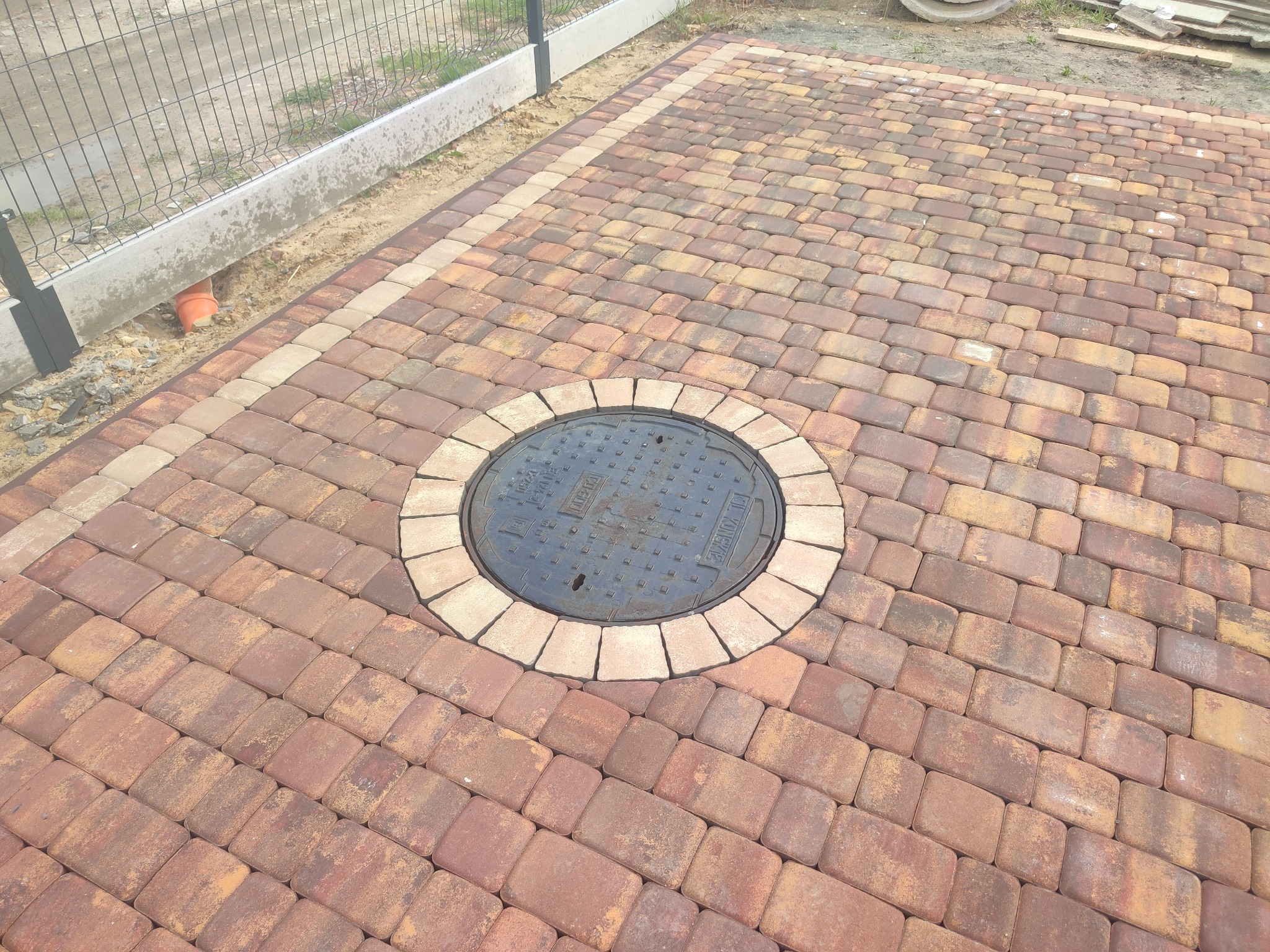 Nowo ułożona, czerwono-brązowa kostka brukowa z dekoracyjnym obramowaniem z jaśniejszych kostek oraz okrągłą studzienką kanalizacyjną na środku, widoczny fragment ogrodzenia panelowego.