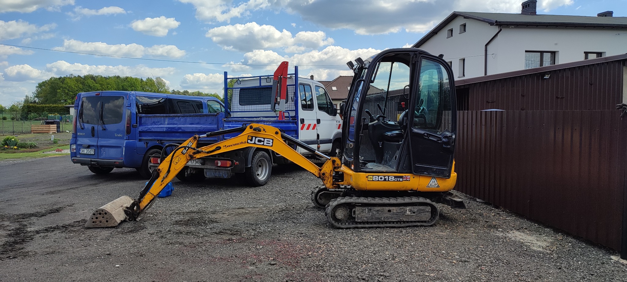 Żółta mini koparka JCB 8018 CTS z łyżką do wykopów, zaparkowana na żwirowym podłożu obok niebieskiego i białego samochodu dostawczego, w tle budynek mieszkalny i błękitne niebo z chmurami.