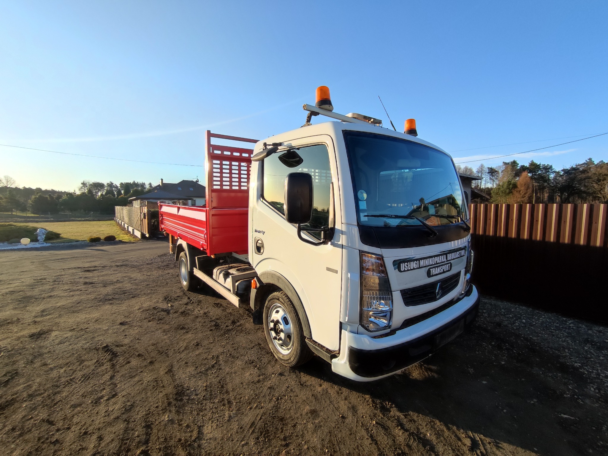Biała ciężarówka z czerwoną skrzynią ładunkową i pomarańczowymi światłami ostrzegawczymi na dachu, zaparkowana na ziemi przy ogrodzeniu i drzewach w słoneczny dzień.