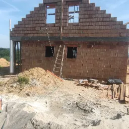 Wznoszony budynek z czerwonej cegły, widoczne otwory okienne, drabina oparta o ścianę, hałda piasku w pierwszym planie, błękitne niebo w tle.