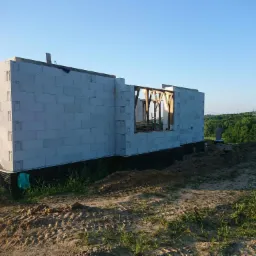 Wznoszone ściany budynku z betonu komórkowego na fundamencie z hydroizolacją, widoczne drewniane nadproża okienne, teren wokół częściowo wyrównany, w tle zieleń drzew.