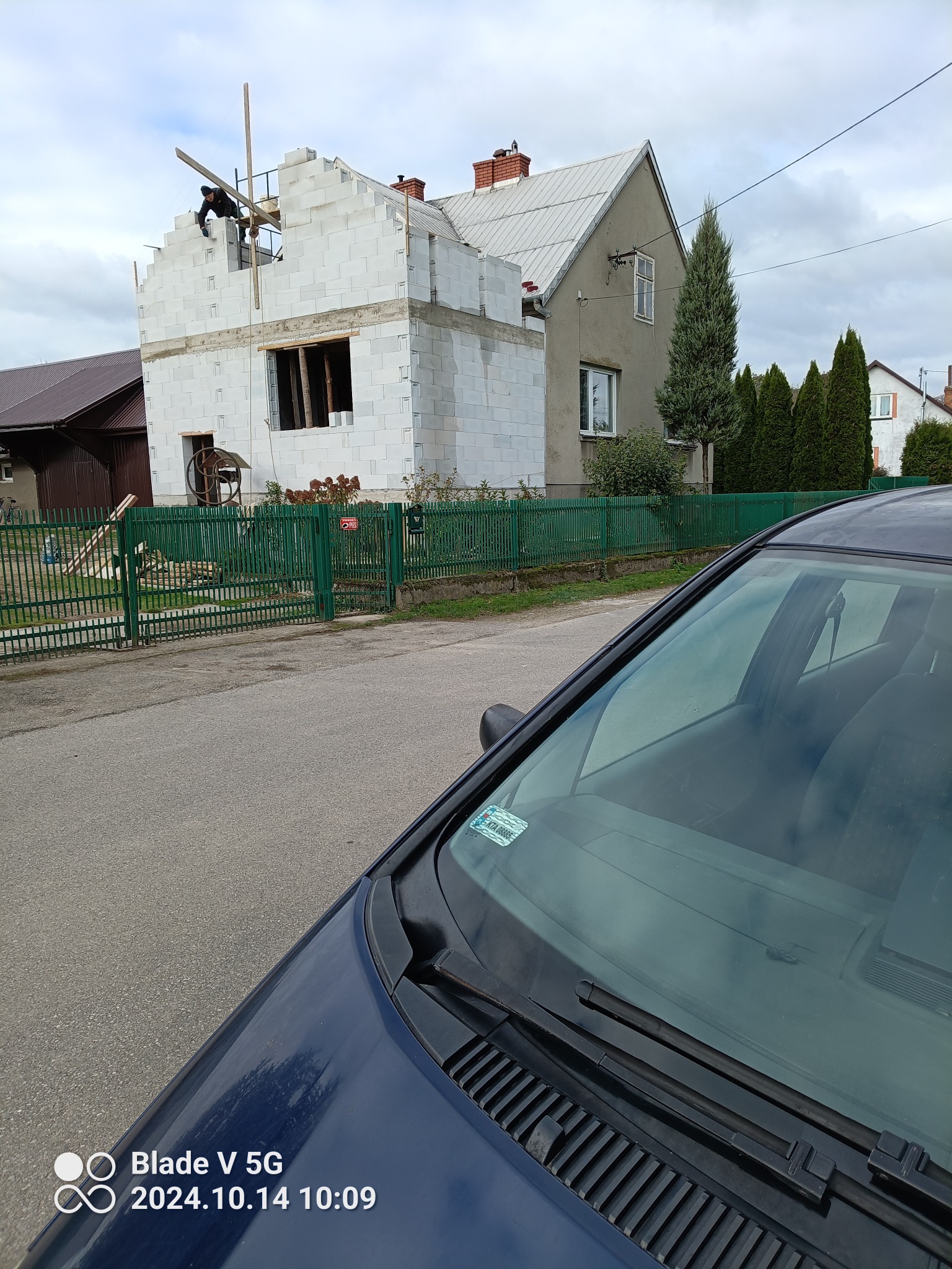 Wznoszenie ścian domu z bloczków białych, widoczny pracownik na rusztowaniu, fragment samochodu na pierwszym planie, w tle zabudowa jednorodzinna.