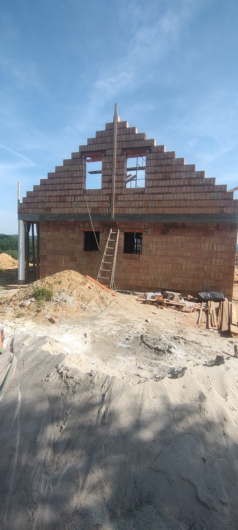 Wznoszony budynek z czerwonej cegły, widoczne otwory okienne, drabina oparta o ścianę, hałda piasku w pierwszym planie, błękitne niebo w tle.