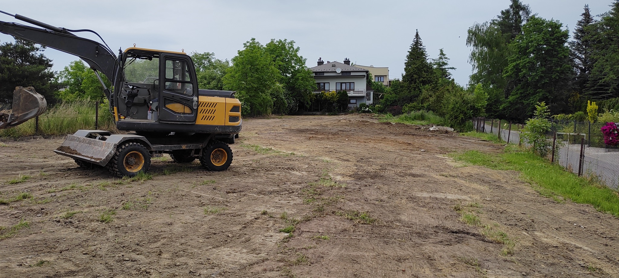 Żółto-czarna koparka kołowa na wyrównanym, przygotowanym pod budowę terenie z widocznym ogrodzeniem i domami w tle.