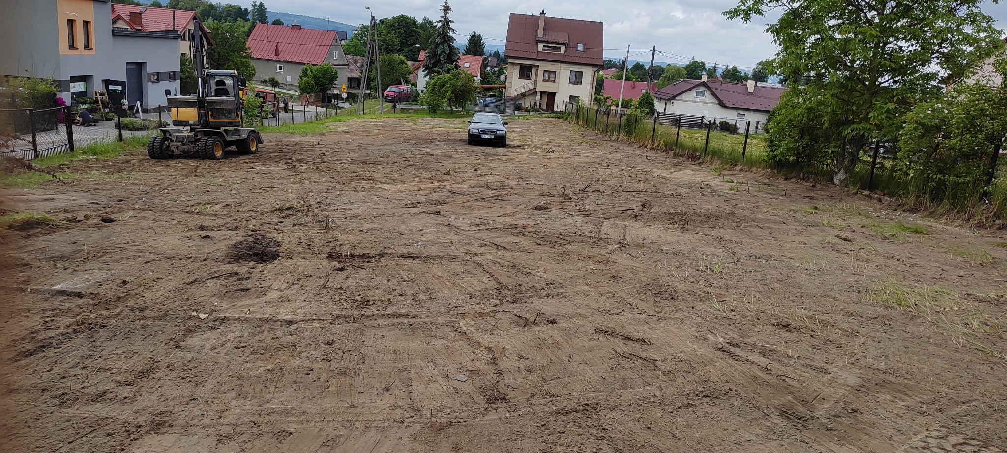 Przygotowany teren pod budowę z widocznymi śladami pracy maszyn ziemnych, na skraju placu stoi koparka kołowa, a w oddali zaparkowany samochód osobowy, w tle zabudowa mieszkalna.
