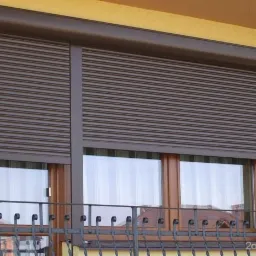 Brązowa roleta zewnętrzna z lamelami, zamontowana na oknie balkonowym z widoczną balustradą, na tle żółtej elewacji budynku.