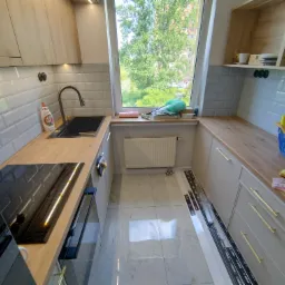 Nowoczesna kuchnia z jasnymi drewnianymi szafkami, szarymi płytkami metra, czarnym zlewem i płytą indukcyjną, widok z okna na zieleń drzew.