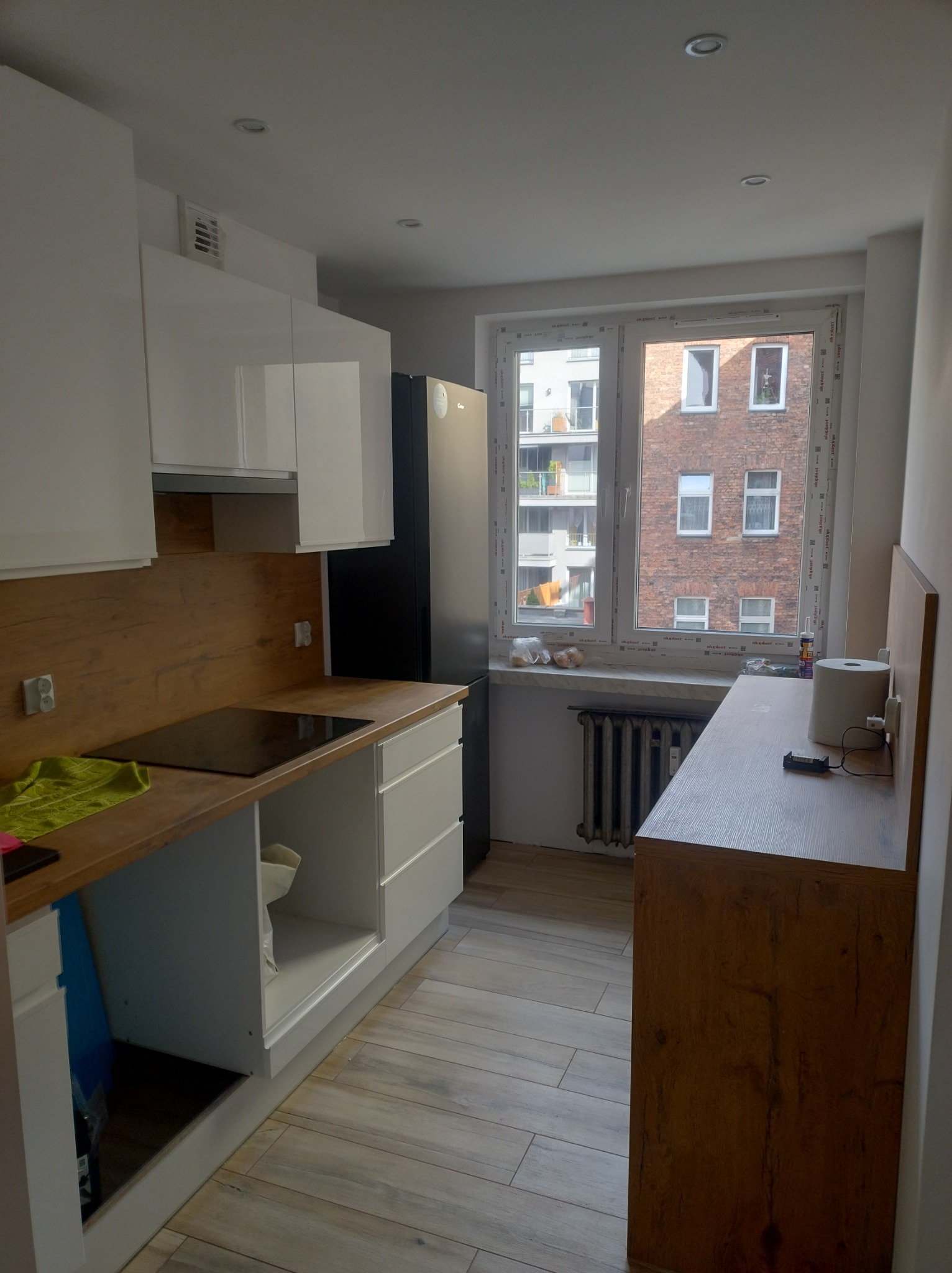 Jasna kuchnia z białymi szafkami i drewnopodobnym blatem, widok na budynek z cegły za oknem, drewniana podłoga.