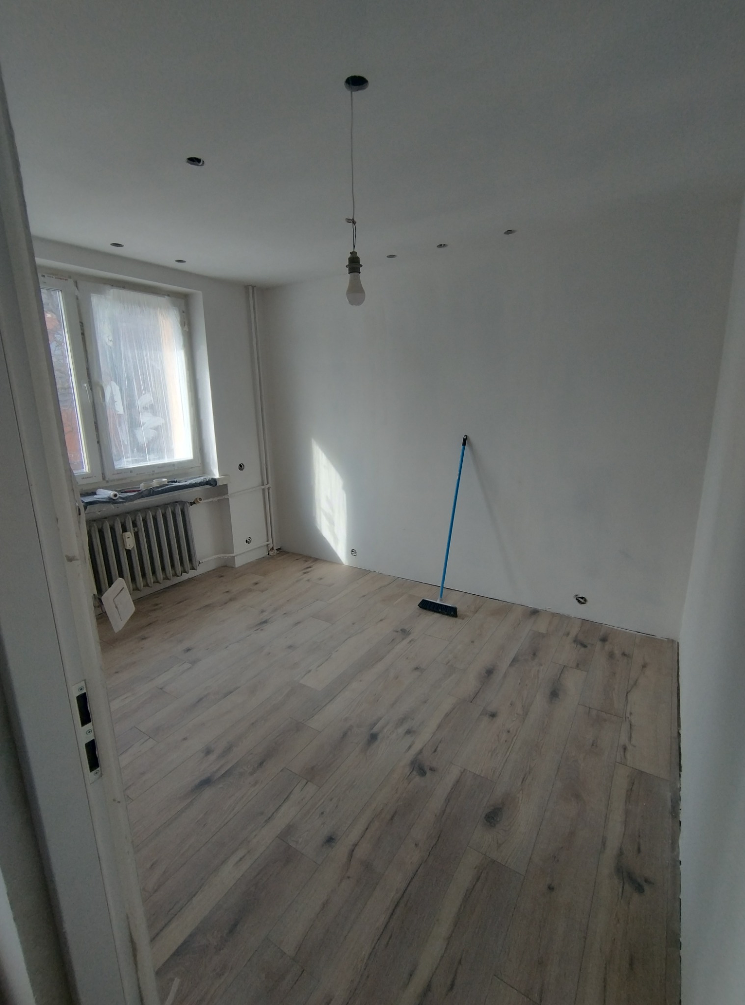 Pusty pokój po remoncie z drewnianą podłogą, świeżo pomalowanymi białymi ścianami, zakrytym oknem i pojedynczą żarówką wiszącą z sufitu, z niebieską zmiotką opartą o ścianę.
